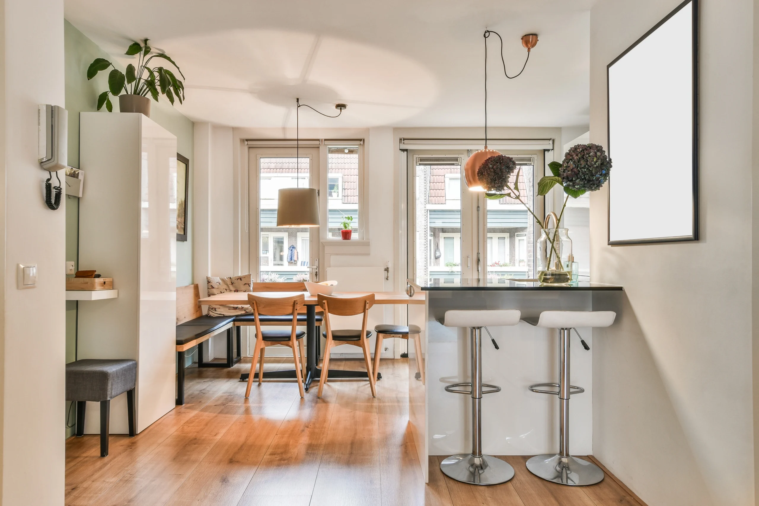 Bright open-plan dining area with a wooden table, corner bench seating, and a modern breakfast bar with two white stools.